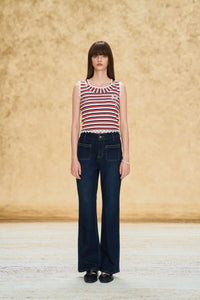 A model stands facing forward wearing a sleeveless top with red, white, and blue horizontal stripes, paired with dark blue wide-leg jeans and black shoes, against a textured beige backdrop.